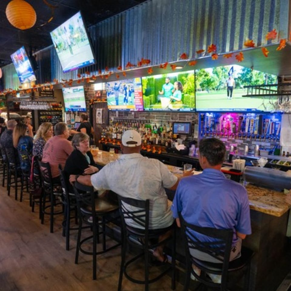 Patrons seated at a lively bar with multiple large screens displaying sports and a wide selection of drinks.